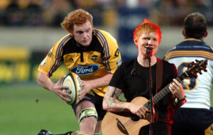 Paul Tito running the ball up the guts, with Ed Sheeran playing guitar in foreground.